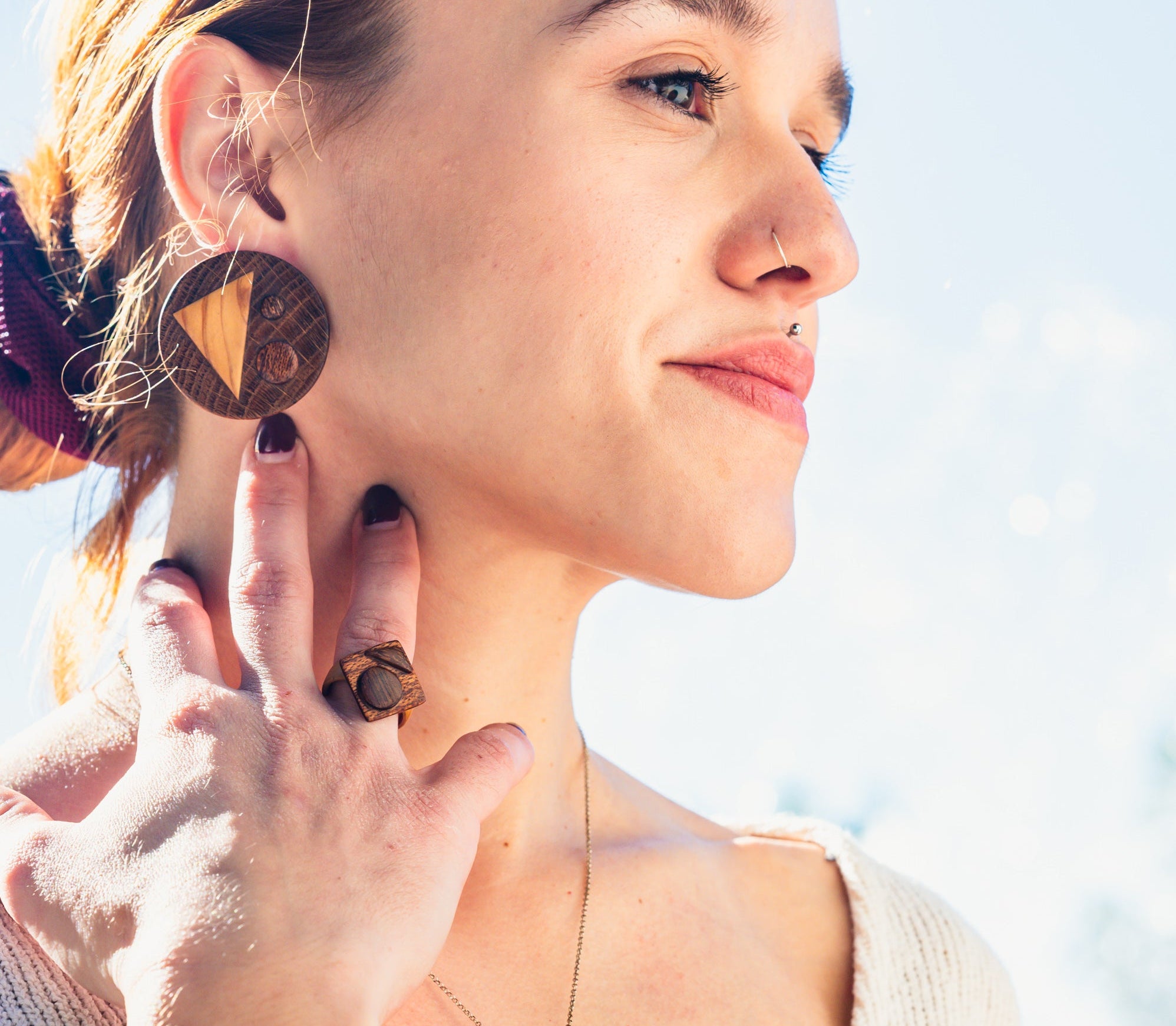 Woman wearing round wooden earrings from four kinds of wood (oak, pine, meranty and iroco), a bended wooden ring that on the top ends square and has also some wooden details. With a blurred outdoor background 