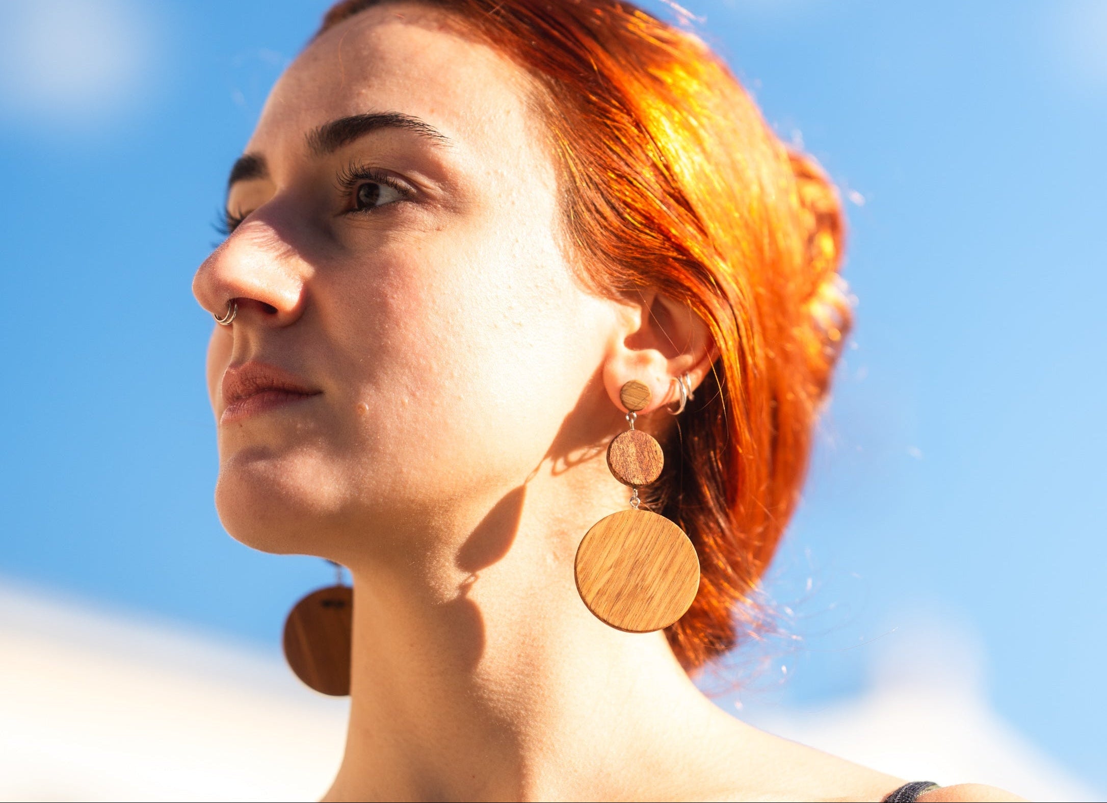 woman with red hair in open space with hangging wooden earrings, three circle pieces connenting with each other. the smaller on the ear and two more under it.